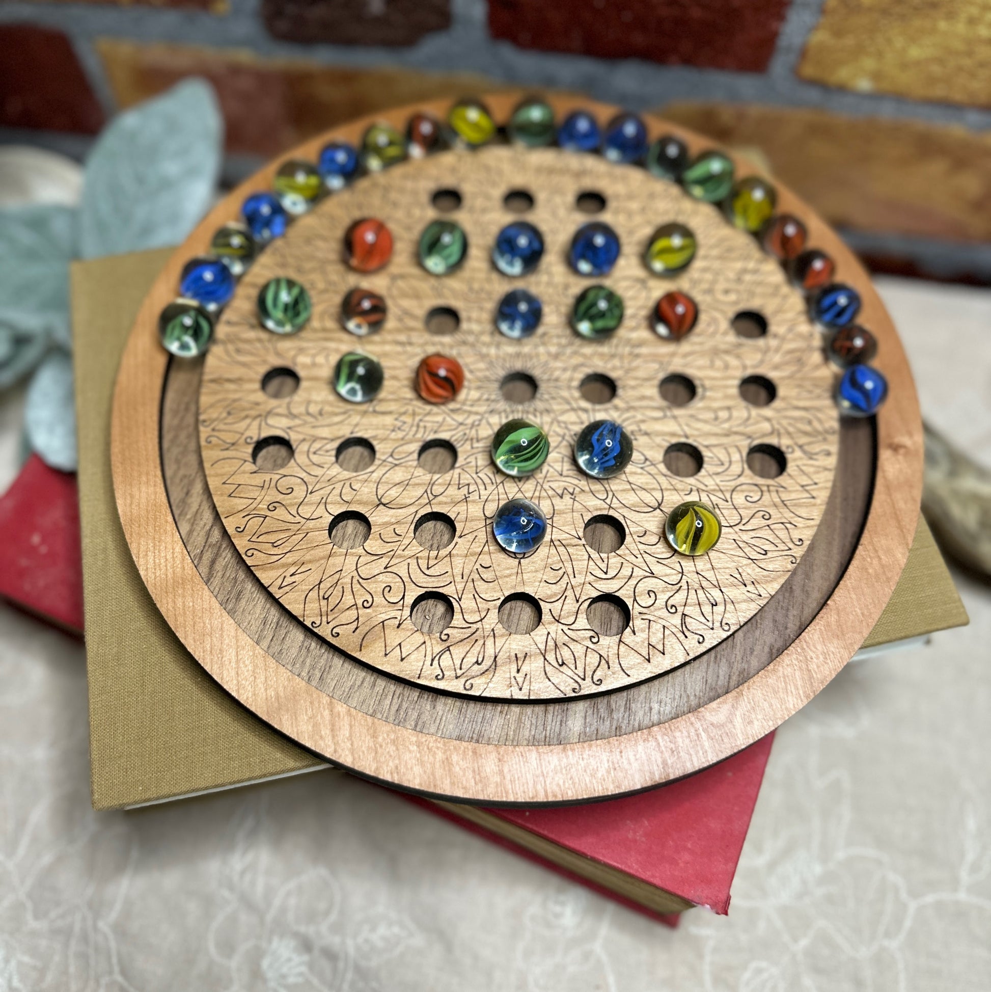 Cherry and Walnut marble solitaire board shown with multi colored marbles. Displayed on a stack of books and ready to be played with. 