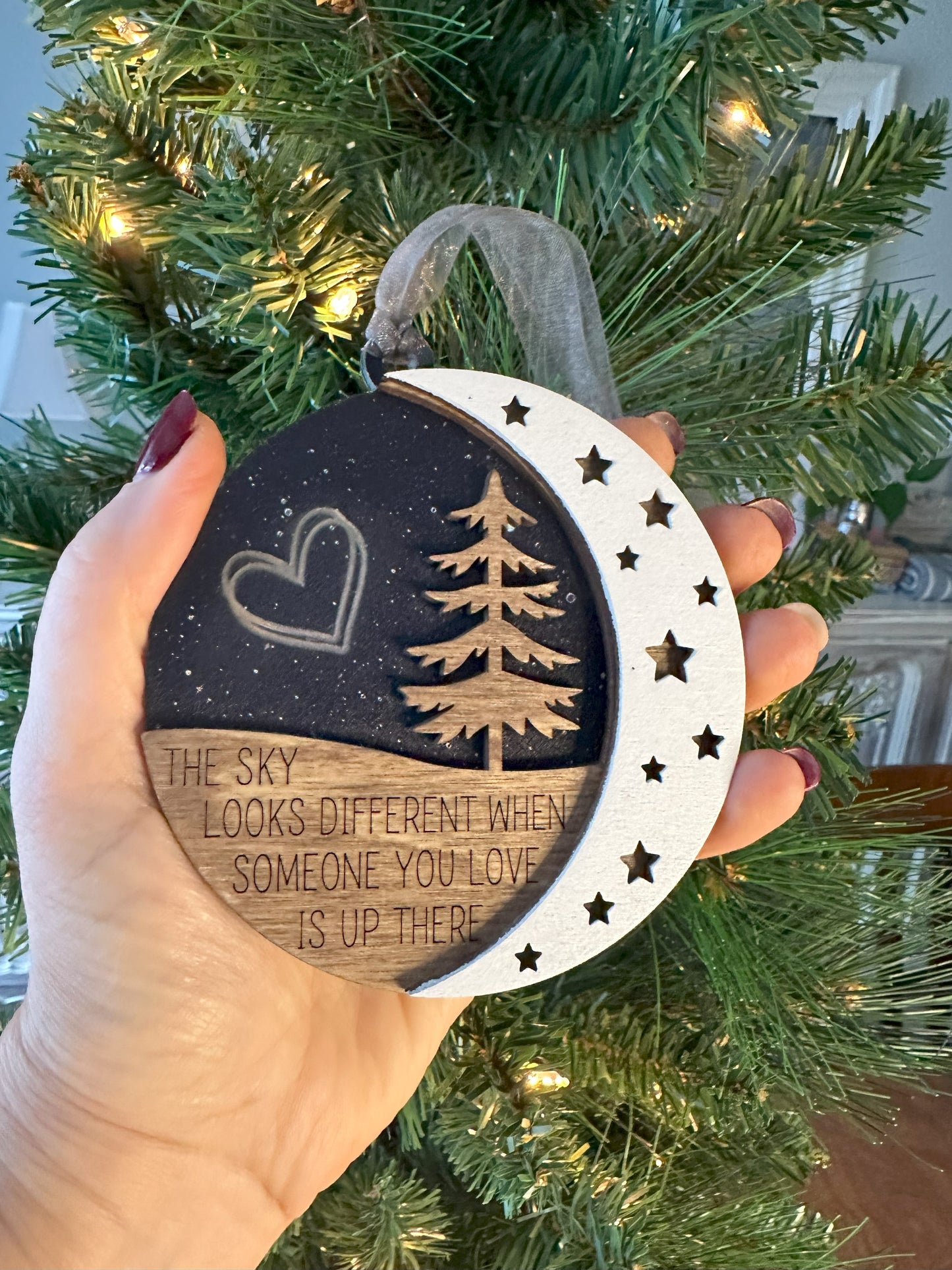 Woman holding ornament for size reference. Three layered ornament that says the sky looks different when someone you love is up there. Black acrylic backer to emulate the evening sky with stars and double heart engraved. Evergreen tree and moon in the for front.