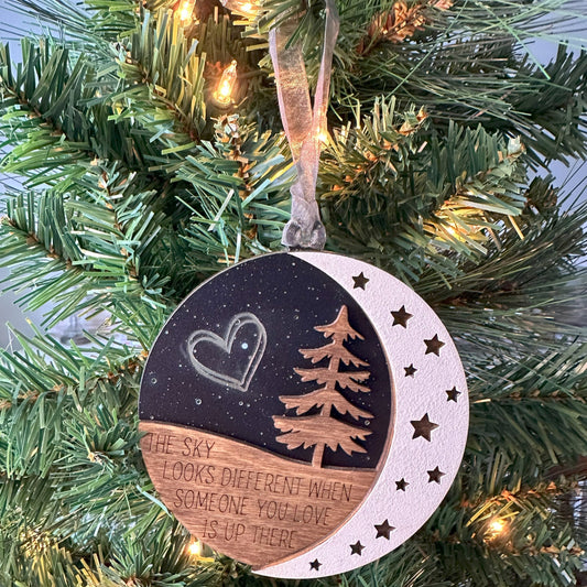 Three layered ornament that says the sky looks different when someone you love is up there. Black acrylic backer to emulate the evening sky with stars and double heart engraved. Evergreen tree and moon in the for front. 