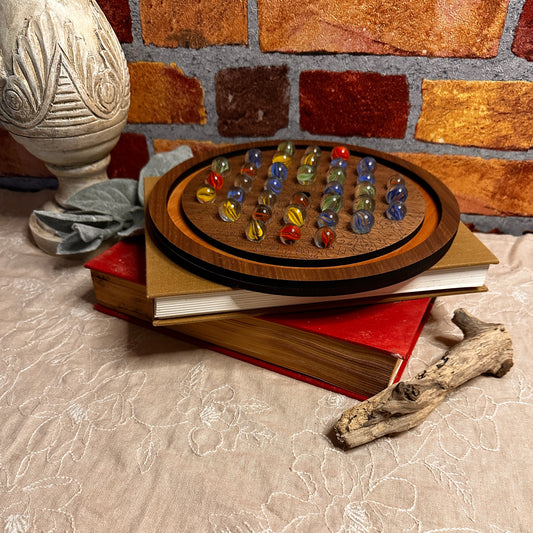 Walnut and Cherry marble solitaire board shown with multi colored marbles. Displayed on a stack of books and ready to be played with. 